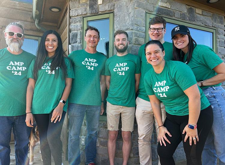Seven Ample team members in matching Amp Camp '24 shirts outside a lodge