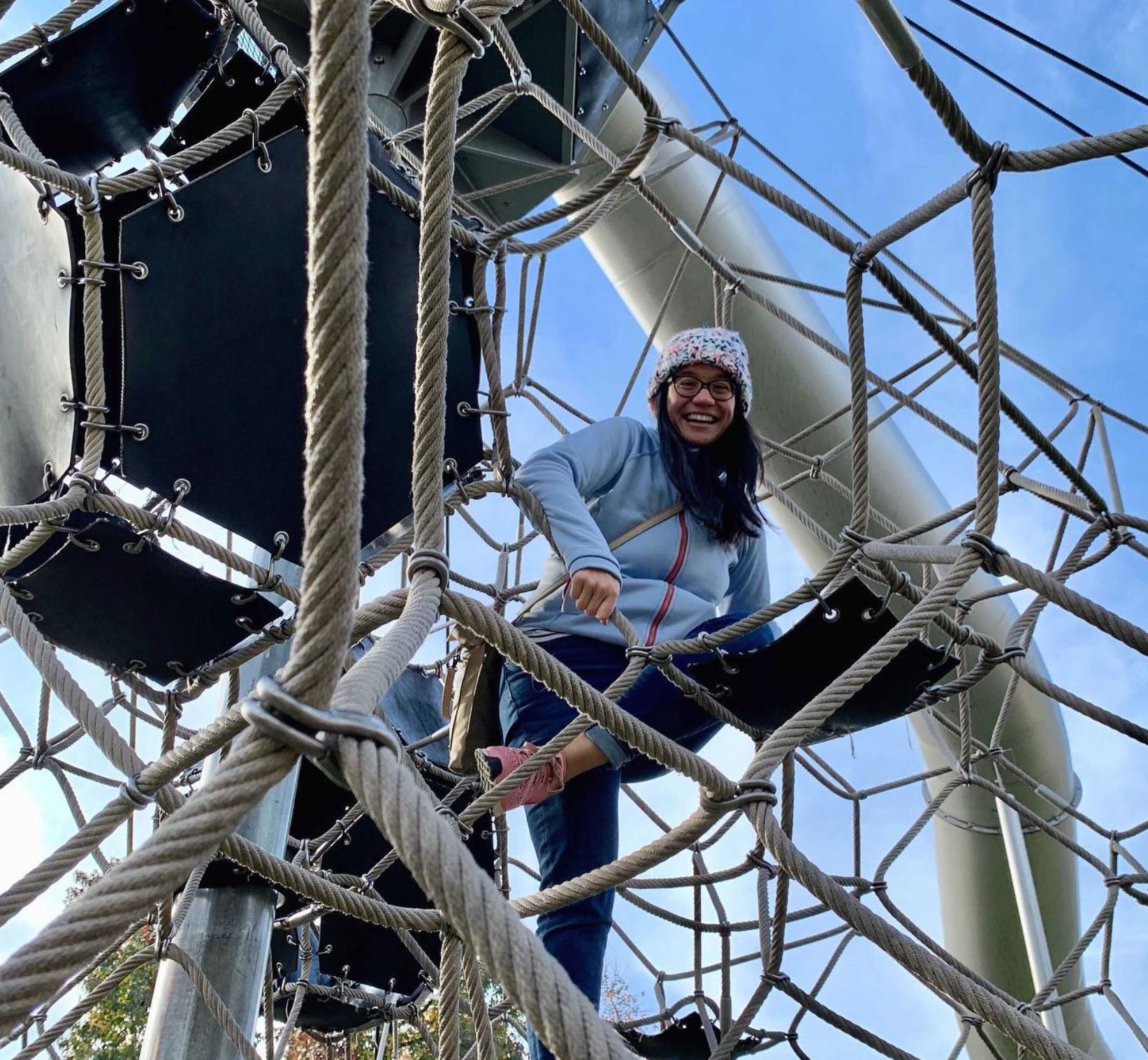 An Ample team member in a knit hat laughing down from high up in a rope climbing net against a blue sky