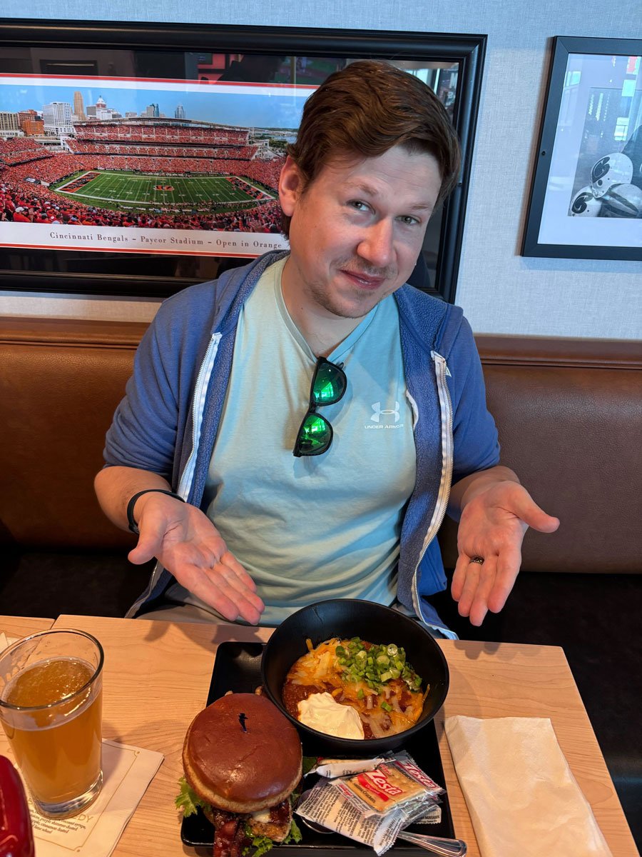 An Ample team member in a restaurant booth presenting a bowl of chili and a sandwich, a Bengals stadium panorama on the wall behind him