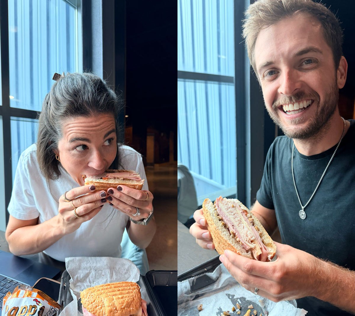Two Ample team members side by side, each digging into a deli sandwich beside a window