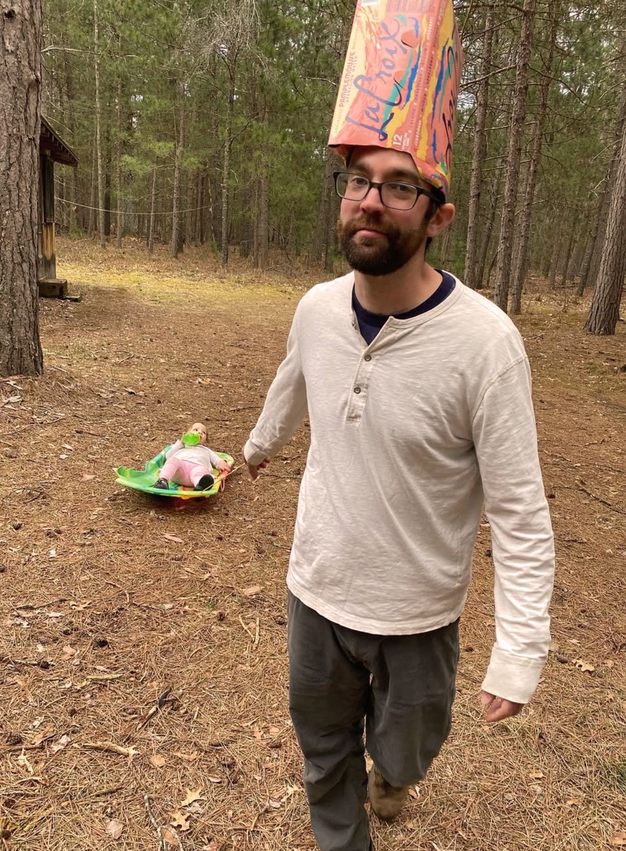An Ample team member in the woods wearing a sparkling-water box as a hat, towing a toddler on a sled behind him